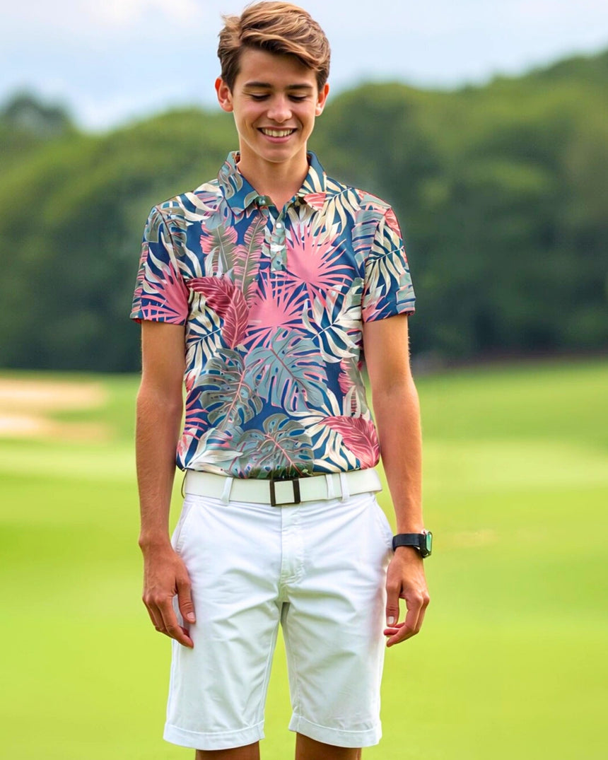 A young man smiles wearing a patterned golf polo, white shorts, and a white belt on a golf course.