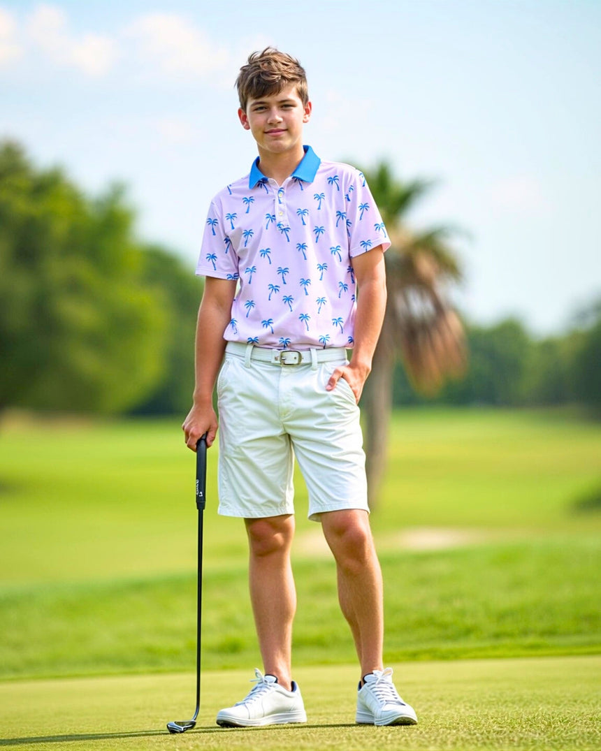 A teenage boy stands on a golf course, wearing a light pink golf polo with a blue palm tree pattern, white shorts, a white belt, and white sneakers, holding a golf club.