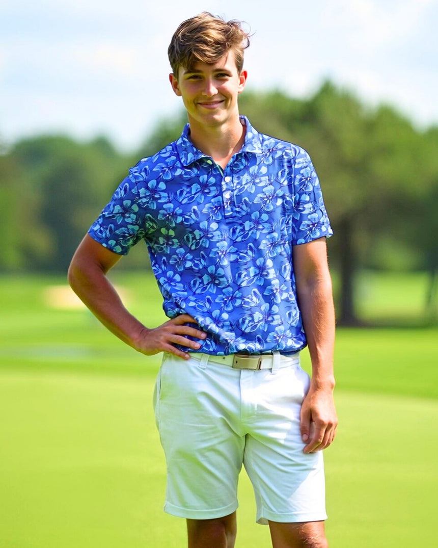 A young man stands on a golf course, wearing a blue floral golf polo and white shorts.