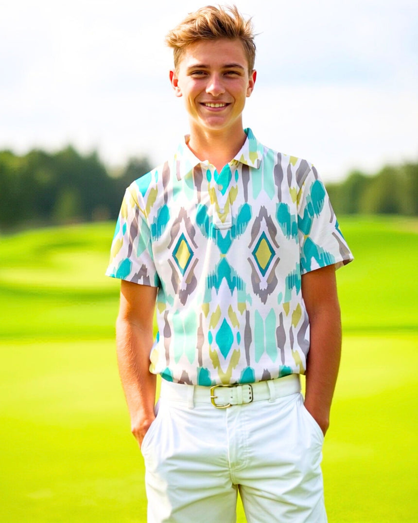 A smiling young man stands on a golf course wearing a patterned golf polo and white pants.