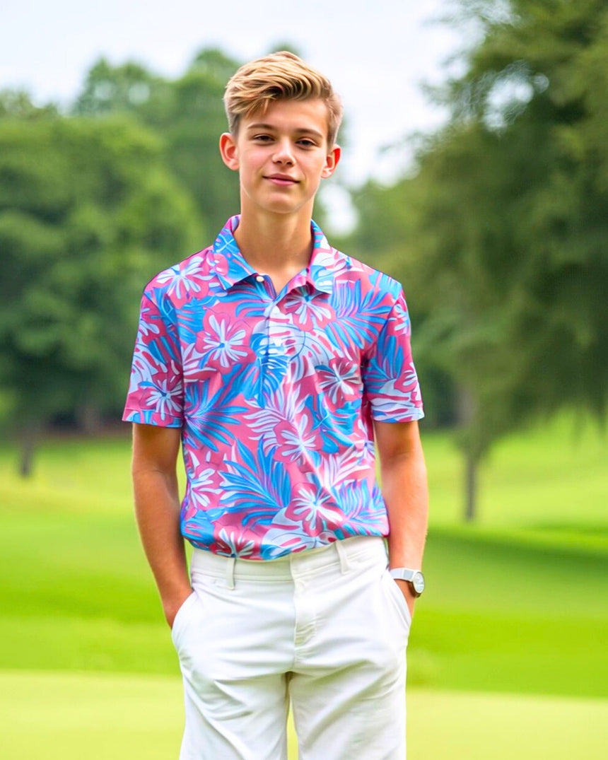 A young man stands on a golf course, wearing a pink and blue floral golf polo and white shorts.