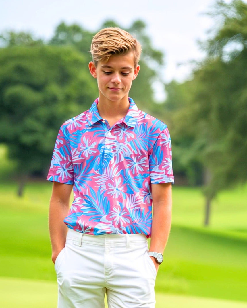 A young man stands on a golf course wearing a pink and blue floral golf polo and white pants.