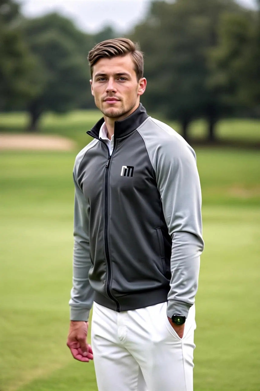 A man wearing a gray and black golf jacket and white pants stands on a golf course.