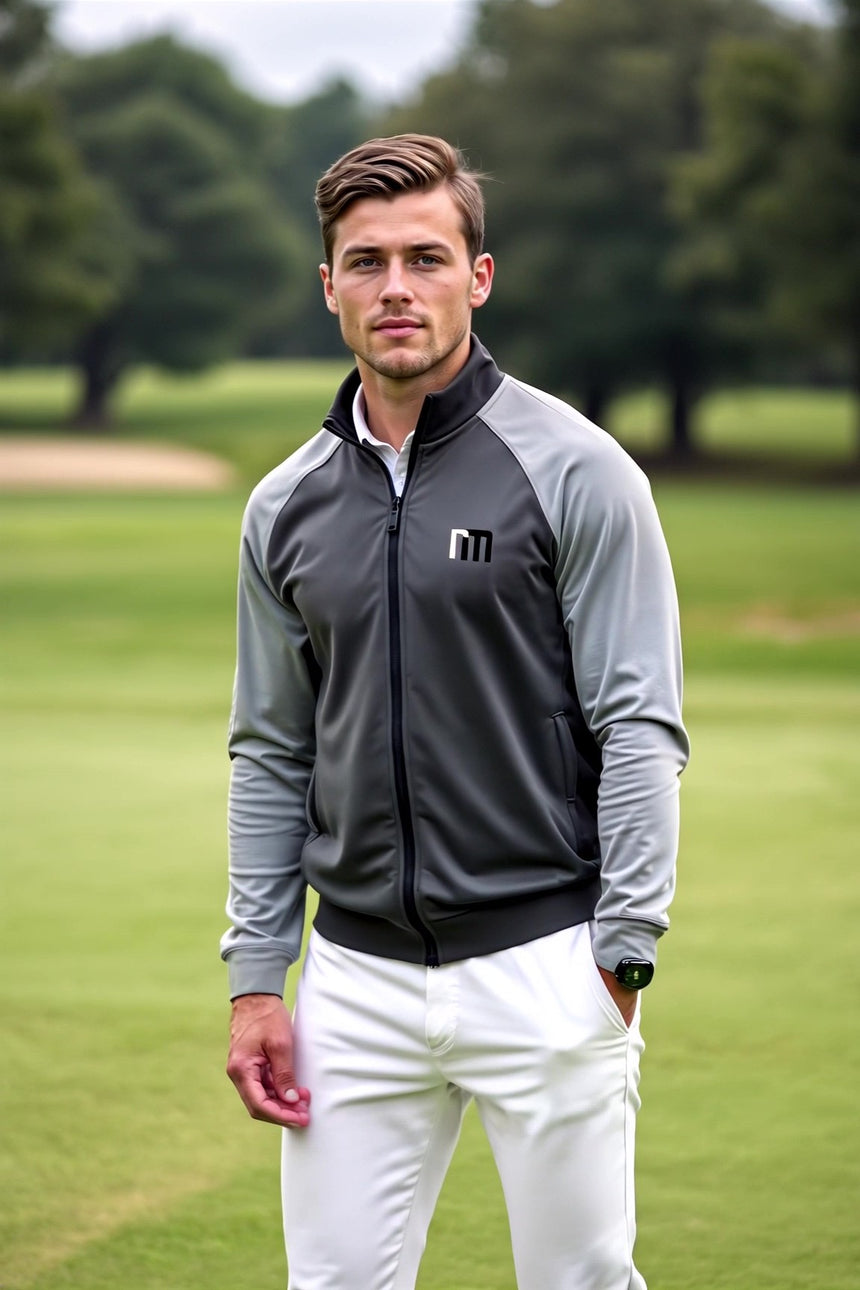 A man stands on a golf course wearing a gray and light gray golf jacket with white pants.