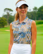 A woman on a golf course smiles while wearing a sleeveless golf polo with a blue, tan, and brown floral pattern, a white visor, and a white skirt.