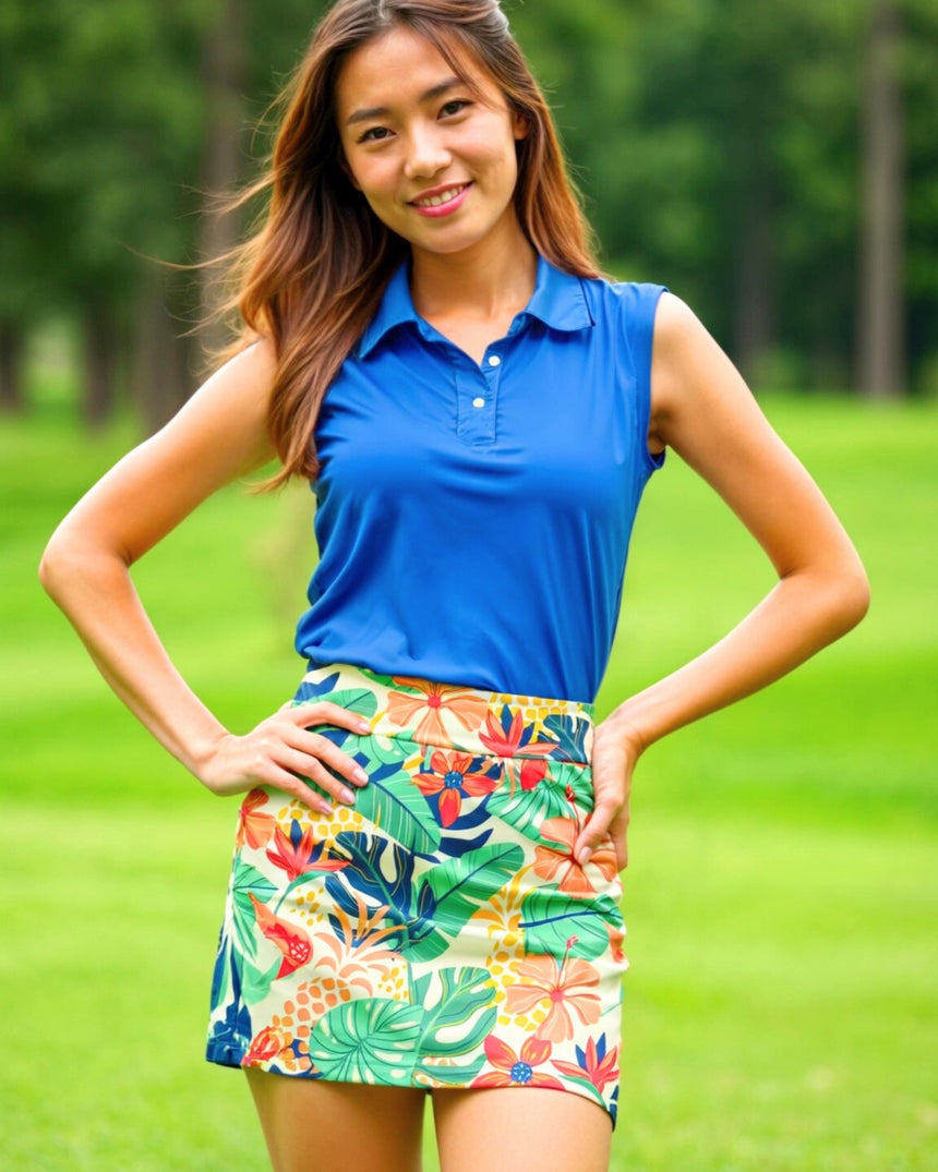A woman in a sleeveless blue golf polo and tropical print golf skirt poses on a golf course.