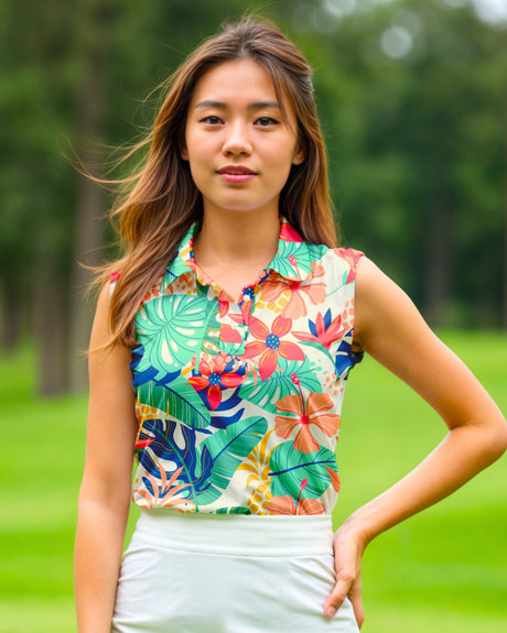 A woman poses on a golf course wearing a sleeveless golf polo with a tropical floral pattern and a white golf skirt.