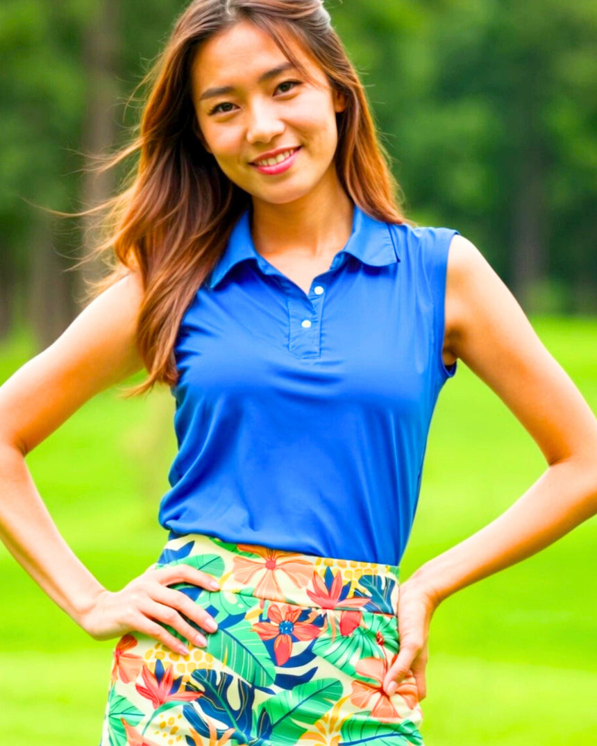 A woman in a sleeveless blue golf polo and floral skirt stands on a golf course.