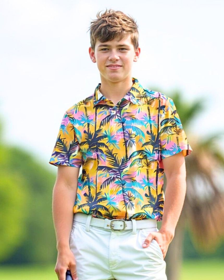 A young man smiles wearing a colorful palm tree golf polo, white shorts, and a white belt on a golf course.