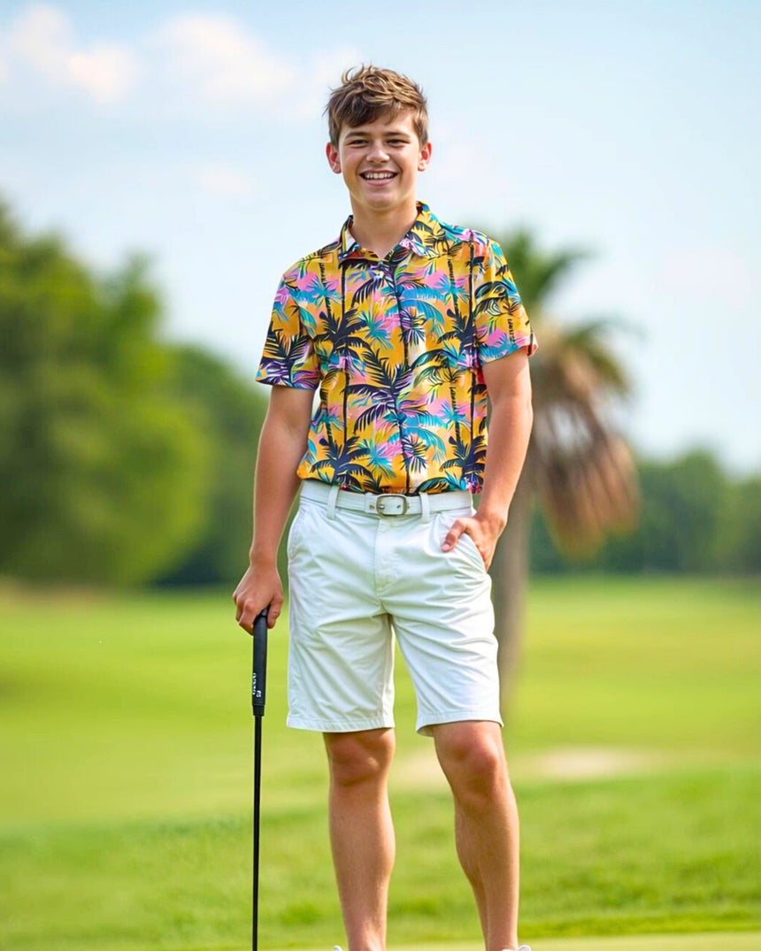 A young man smiles on a golf course, wearing a colorful palm tree print golf polo, white shorts, and holding a putter.
