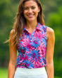 A smiling woman in a pink sleeveless golf polo with a blue and purple floral pattern and a white golf skirt stands against a green background.