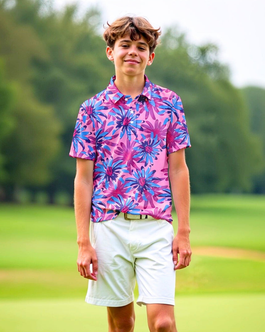 A young man stands on a golf course, wearing a pink golf polo with a blue and purple floral pattern and white shorts.