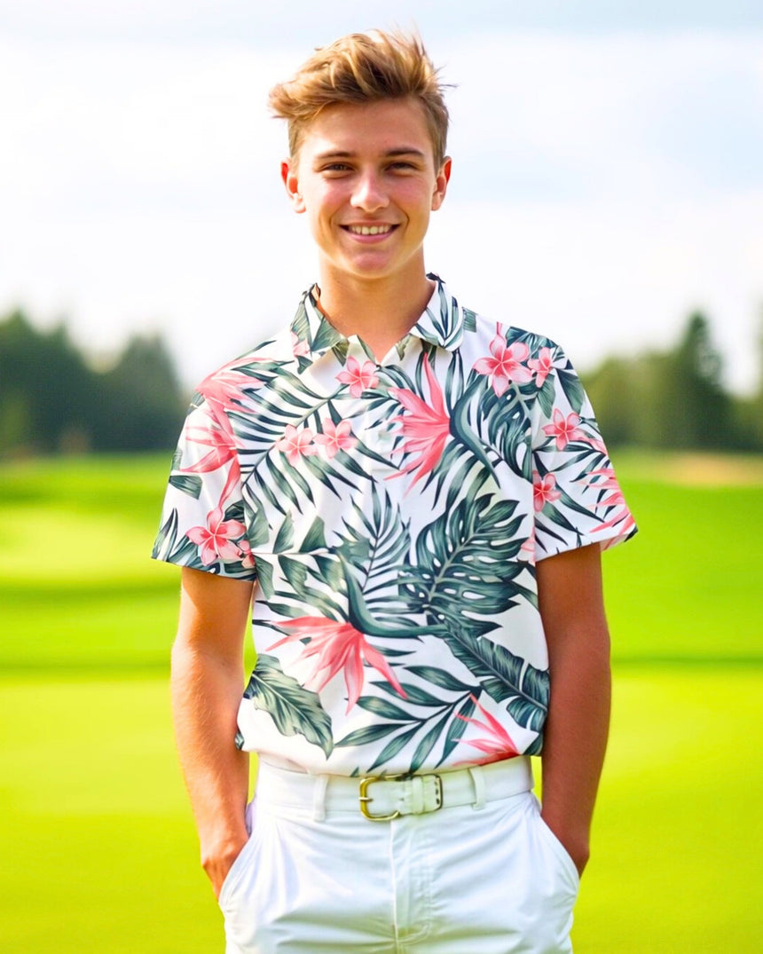 A smiling young man stands on a golf course, wearing a white golf polo with a tropical floral pattern, white pants, and a white belt.