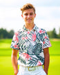 A smiling young man in a floral golf polo and white pants stands on a golf course.