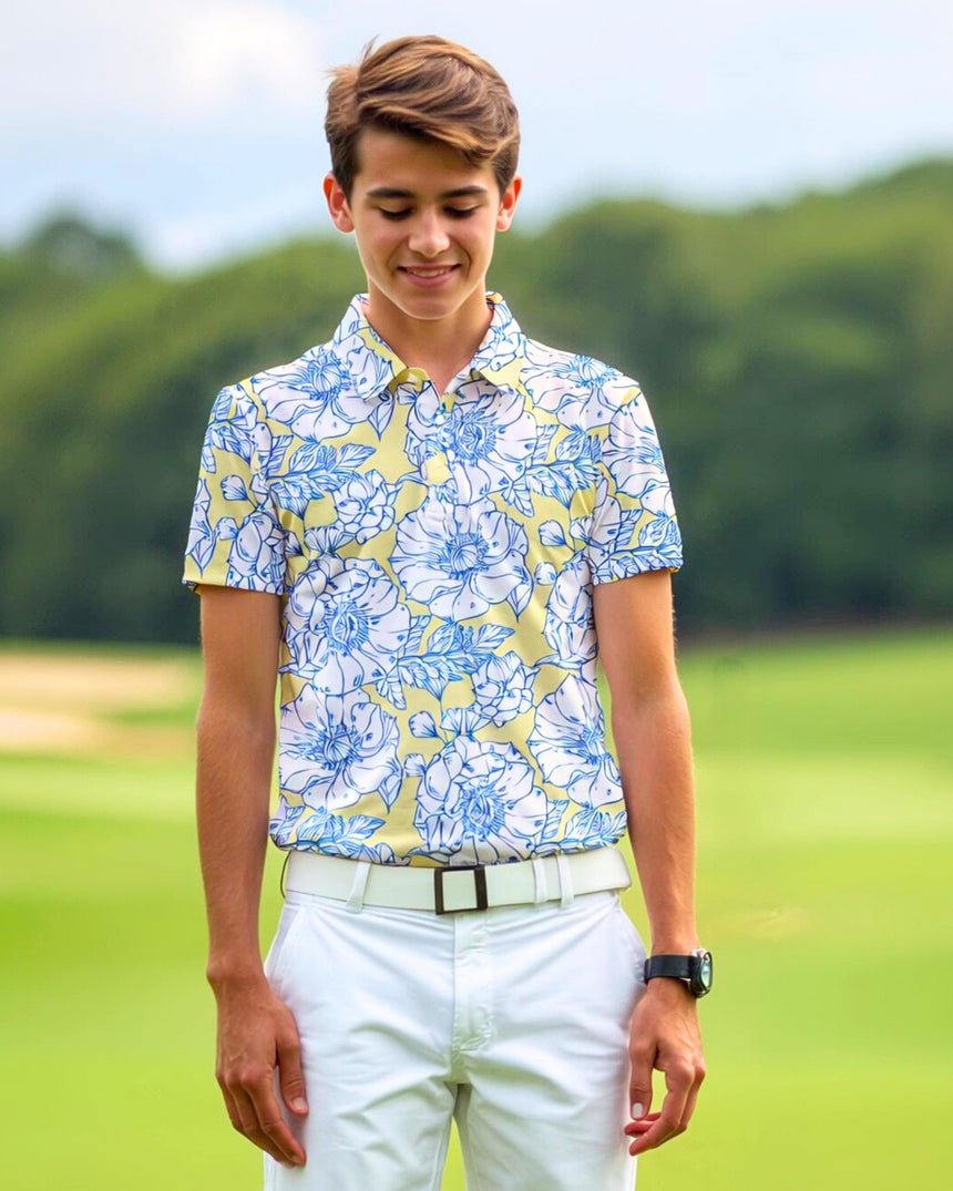 A young man wearing a floral golf polo, white pants, and a white belt stands on a golf course.