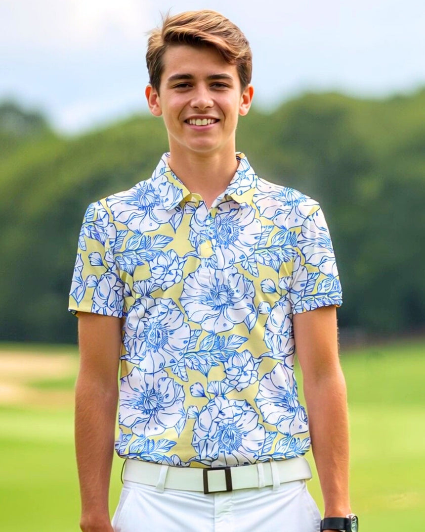 A smiling young man stands on a golf course wearing a yellow golf polo with a blue and white floral pattern, white pants, and a white belt.