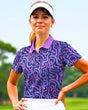 A woman in a purple patterned golf polo and white visor stands on a golf course.