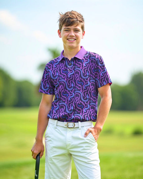 A smiling young man stands on a golf course, wearing a purple patterned golf polo, white shorts, and a white belt. He holds a golf club and has one hand in his pocket.