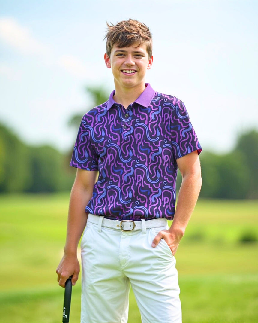 A smiling young man stands on a golf course, wearing a purple patterned golf polo, white shorts, and a white belt. He holds a golf club and has one hand in his pocket.