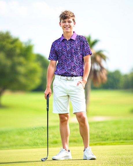 A young man smiles while standing on a golf course, wearing a purple patterned golf polo, white shorts, and white sneakers, holding a golf club.
