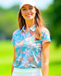 A woman smiles wearing a floral golf polo, white skirt, and white hat.
