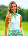 A blonde woman smiles at the camera wearing a sleeveless white golf polo with a mint green collar and placket, paired with a floral print golf skirt.
