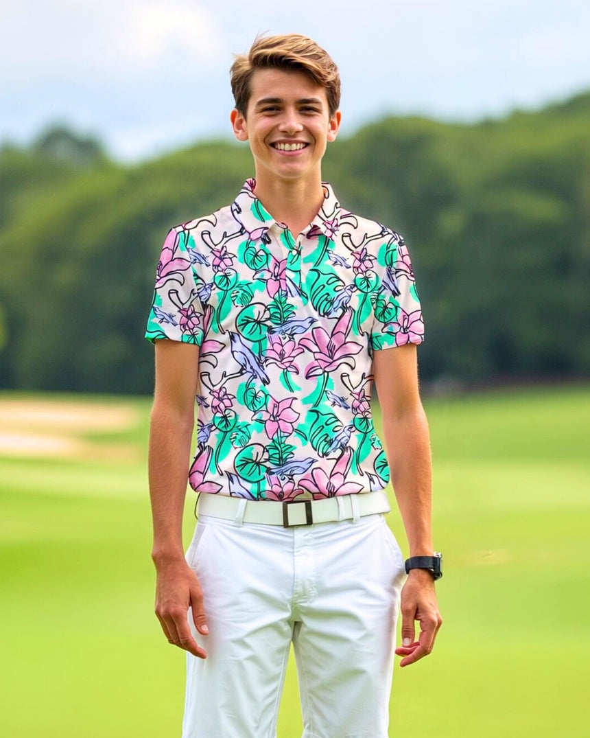 A young man smiles while wearing a golf polo with a floral and bird pattern, white pants, and a white belt on a golf course.