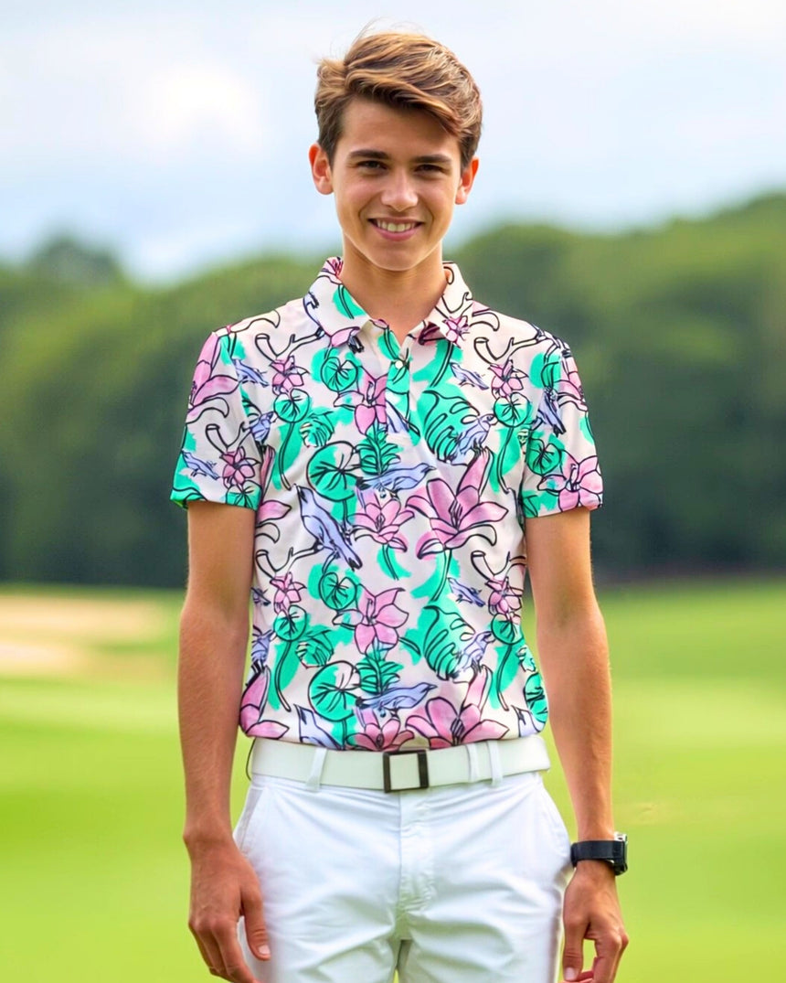 A smiling young man stands on a golf course wearing a white golf outfit and a colorful floral golf polo.