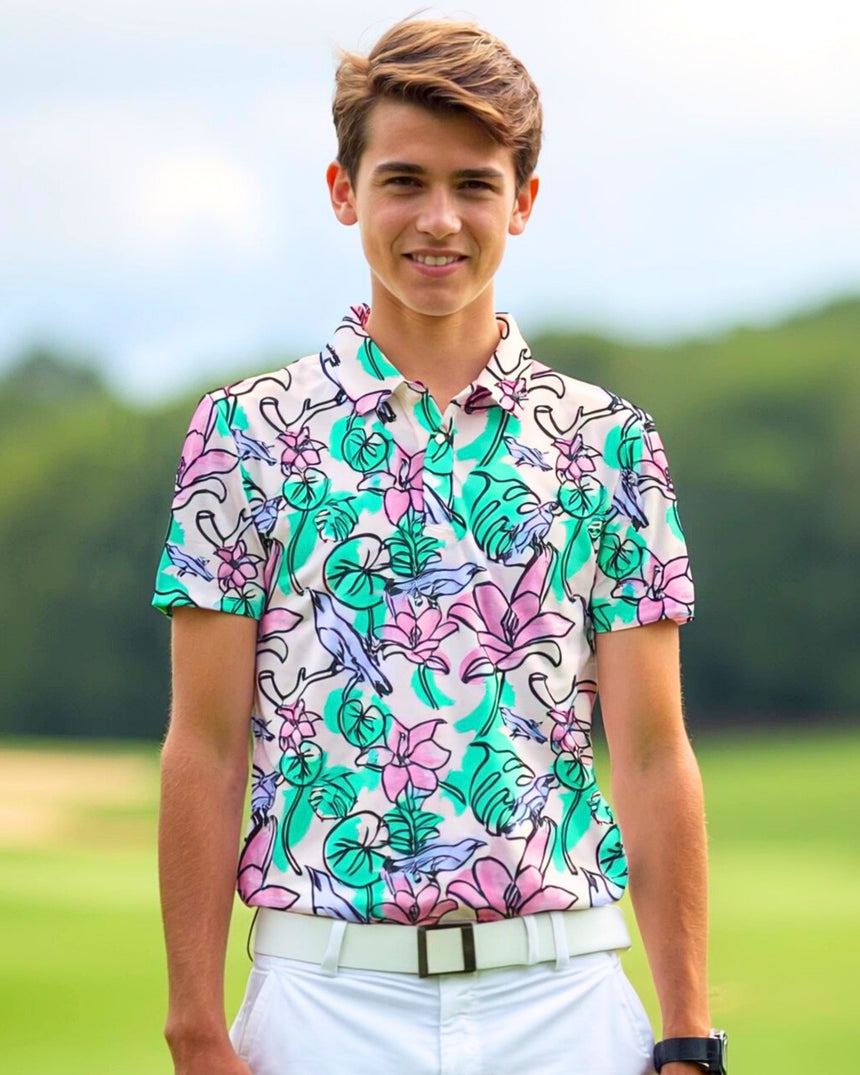 A smiling young man in a floral golf polo, white pants, and a white belt stands on a golf course.