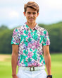 A smiling young man in a floral golf polo, white pants, and a white belt stands on a golf course.