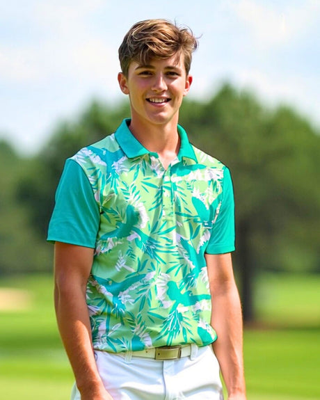 A young man smiles wearing a green and white floral golf polo, white pants, and a white belt, standing on a golf course.