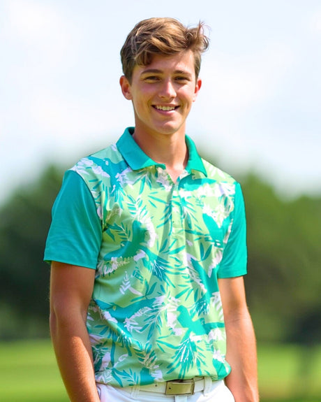 A young man smiles, wearing a golf polo with a light green and turquoise tropical print, paired with white pants.