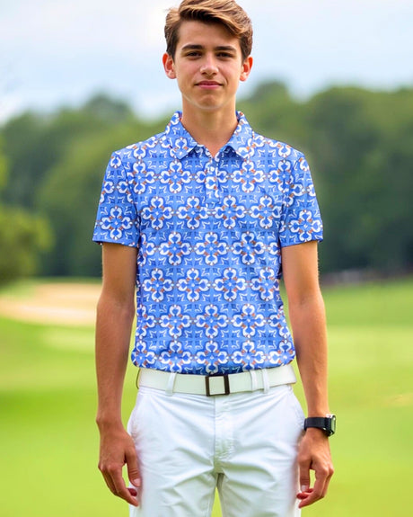 A young man stands on a golf course wearing a blue patterned golf polo, white pants, and a white belt.