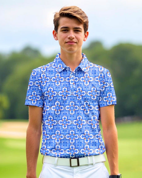 A young man wearing a blue patterned golf polo shirt, white pants, and a white belt stands on a golf course.