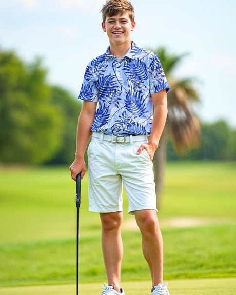A young man smiles while wearing a blue leaf-patterned golf polo, white shorts, and white sneakers on a golf course. He holds a golf club and has one hand in his pocket.