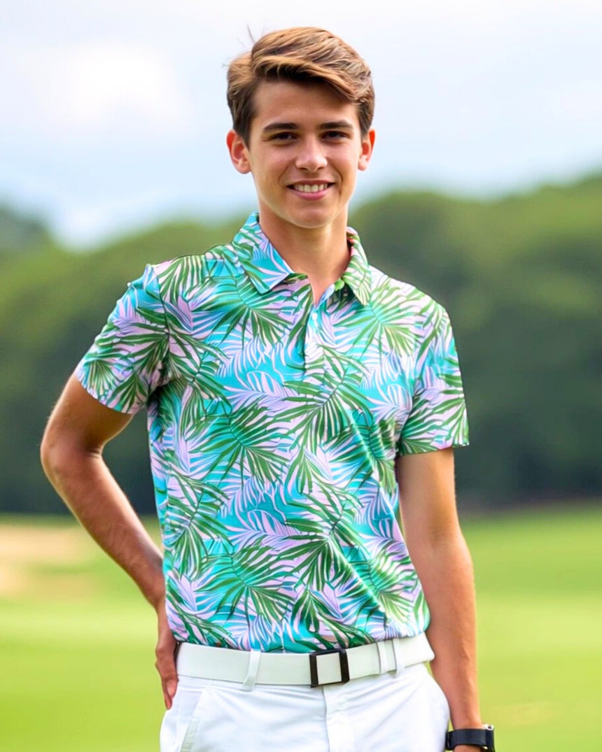 A young man smiles wearing a golf polo with a tropical leaf pattern in shades of green, blue, and lavender, paired with white pants and a white belt, standing on a golf course.