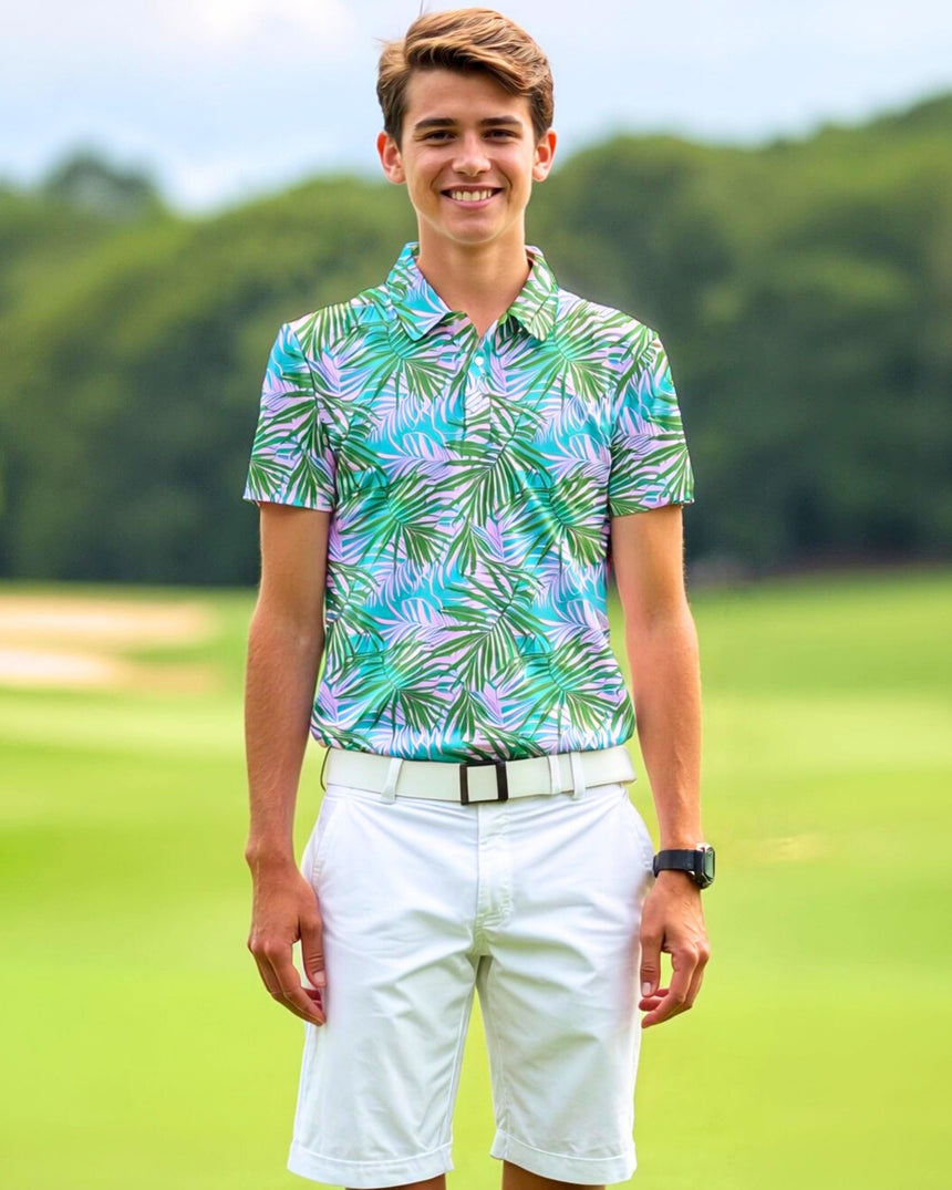 A smiling young man stands on a golf course wearing a golf polo with a tropical leaf pattern in shades of green, blue, and lavender, paired with white shorts and a white belt.