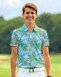 A smiling young man stands on a golf course wearing a short-sleeved golf polo with a tropical leaf pattern in shades of green, blue, and lavender, paired with white pants and a white belt.