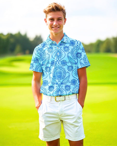 A young man smiles wearing a patterned blue golf polo, white shorts, and a white belt on a golf course.