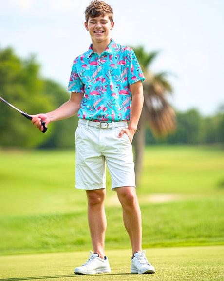 A young man stands on a golf course, smiling and holding a golf club. He is wearing a bright blue golf polo with a flamingo pattern, white shorts, a white belt, and white golf shoes.
