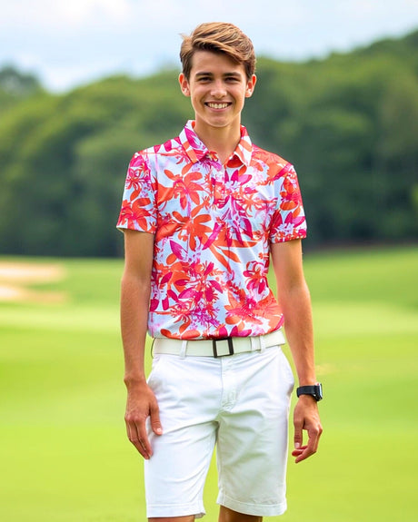 A smiling young man stands on a golf course wearing a pink and orange floral golf polo, white shorts, and a white belt.