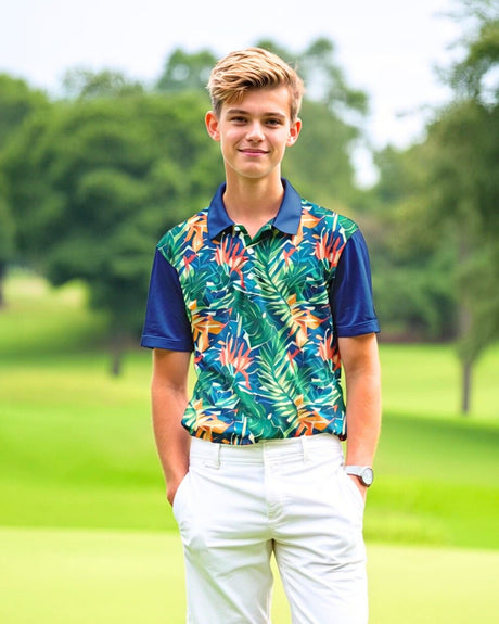 A young man smiles wearing a golf polo with a tropical print and navy blue sleeves, paired with white golf pants, standing on a golf course.