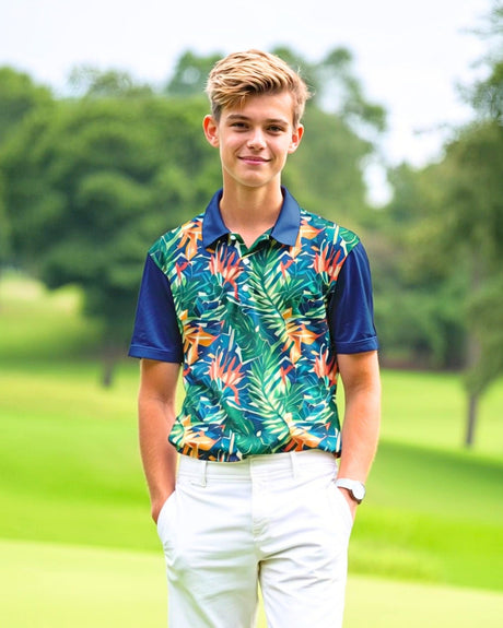 A young man stands on a golf course, wearing a golf polo with a tropical print and white pants.
