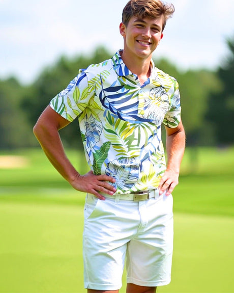 A smiling young man stands on a golf course, wearing a short-sleeved golf polo with a vibrant tropical print of green, yellow, and blue leaves and white golf shorts. He has his hands on his hips.