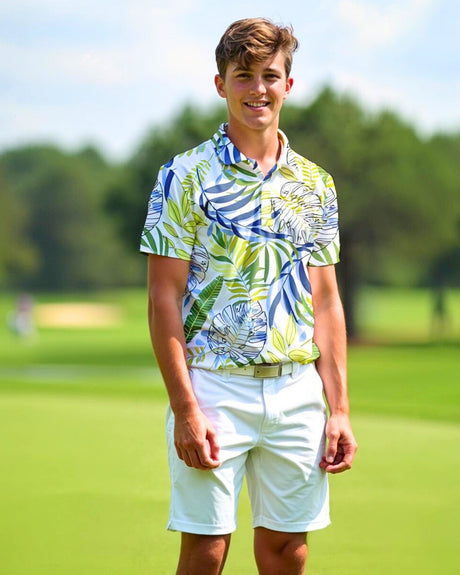 A young man stands smiling on a golf course, wearing a white polo shirt with a blue, green, and yellow tropical leaf pattern and white shorts.