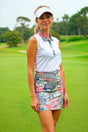 A woman in a white sleeveless golf polo and tropical print skirt stands on a golf course.