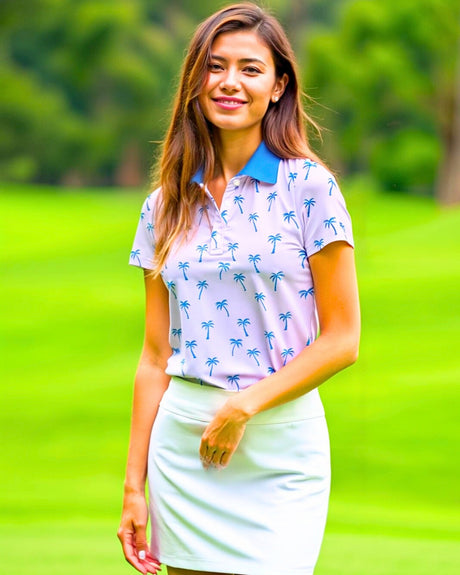 A woman smiles wearing a pink golf polo with blue palm trees and a white skirt on a golf course.