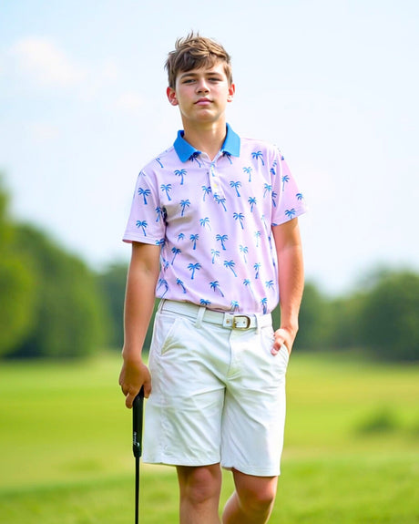 A young man stands on a golf course wearing a light pink golf polo with blue palm trees, white shorts, and holding a golf club.