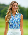 A woman smiles wearing a sleeveless golf polo with a blue, green, and white geometric floral pattern and a white golf skirt.