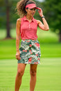 A woman in a sleeveless coral golf polo and floral skirt stands on a golf course.